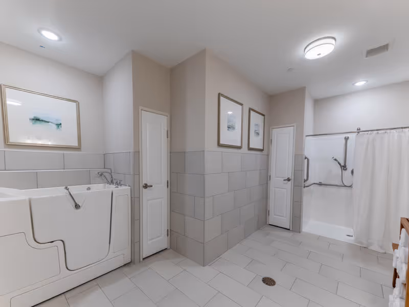A spacious, accessible bathroom featuring a walk-in bathtub with a door on the left and a walk-in shower with grab bars and a curtain on the right. The walls are tiled halfway up with light gray tiles and painted beige above. There are two white doors in the center wall, three framed pictures hanging above the tiles, and a wooden towel rack with white towels on the right side. The floor is tiled with large light gray tiles, and the ceiling has recessed lighting.