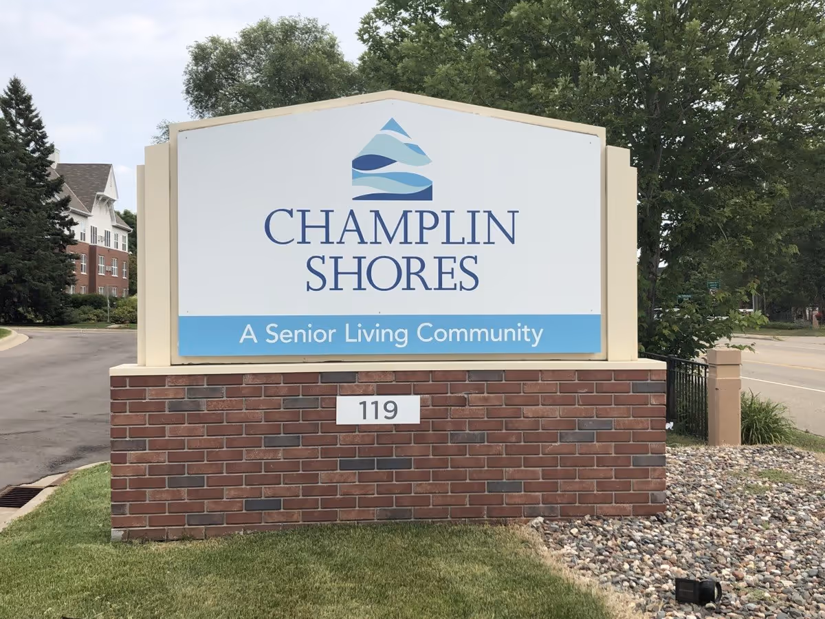 Outdoor sign for Champlin Shores, a senior living community, mounted on a brick base with the address number 119. The sign features a blue and white logo above the community name. Trees, a road, and part of a building are visible in the background.
