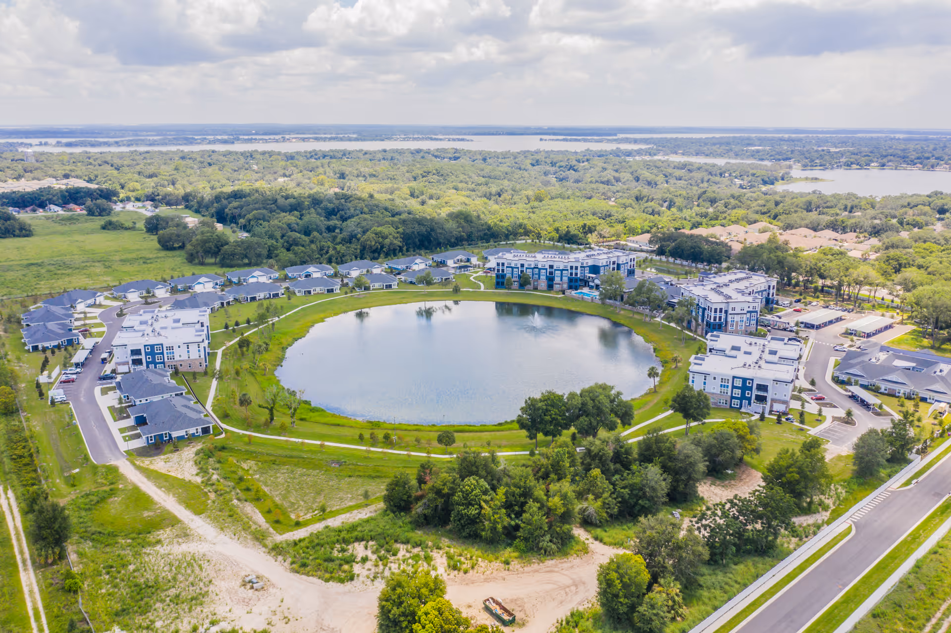 Aerial view of Waterman Village Information Center showing a large circular pond with a fountain in the center, surrounded by multiple residential buildings and greenery. Roads and parking areas are visible around the buildings, with a backdrop of dense trees and distant water bodies under a partly cloudy sky.