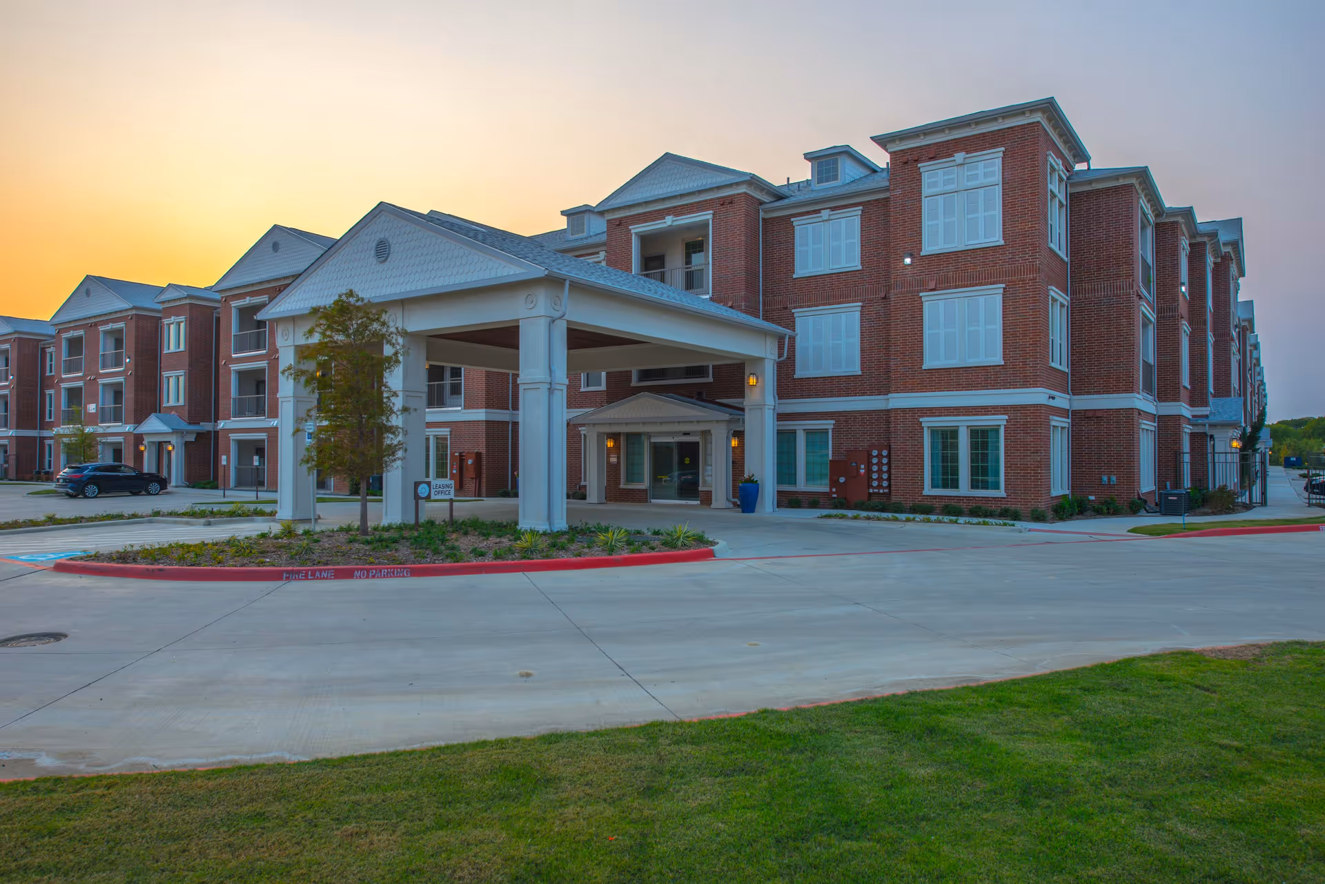 Front entrance of a three-story red brick senior living building with a covered drive-through portico at sunset.
