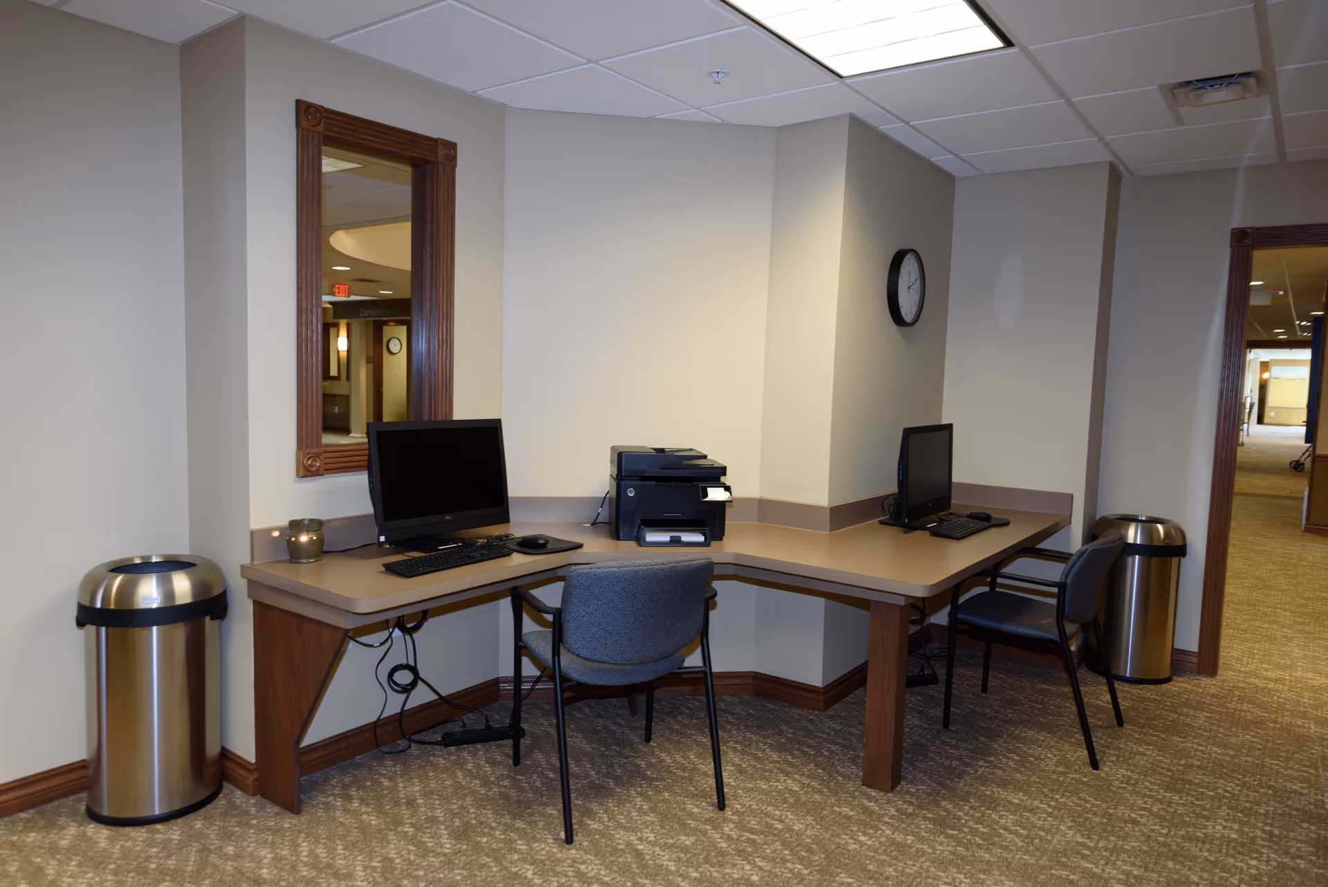 A small office area in a retirement facility with two computer workstations on an L-shaped desk, each with a chair. There is a printer between the two computers, a wall clock, two stainless steel trash bins, and a mirror on the wall. The area has beige walls and carpeted flooring.