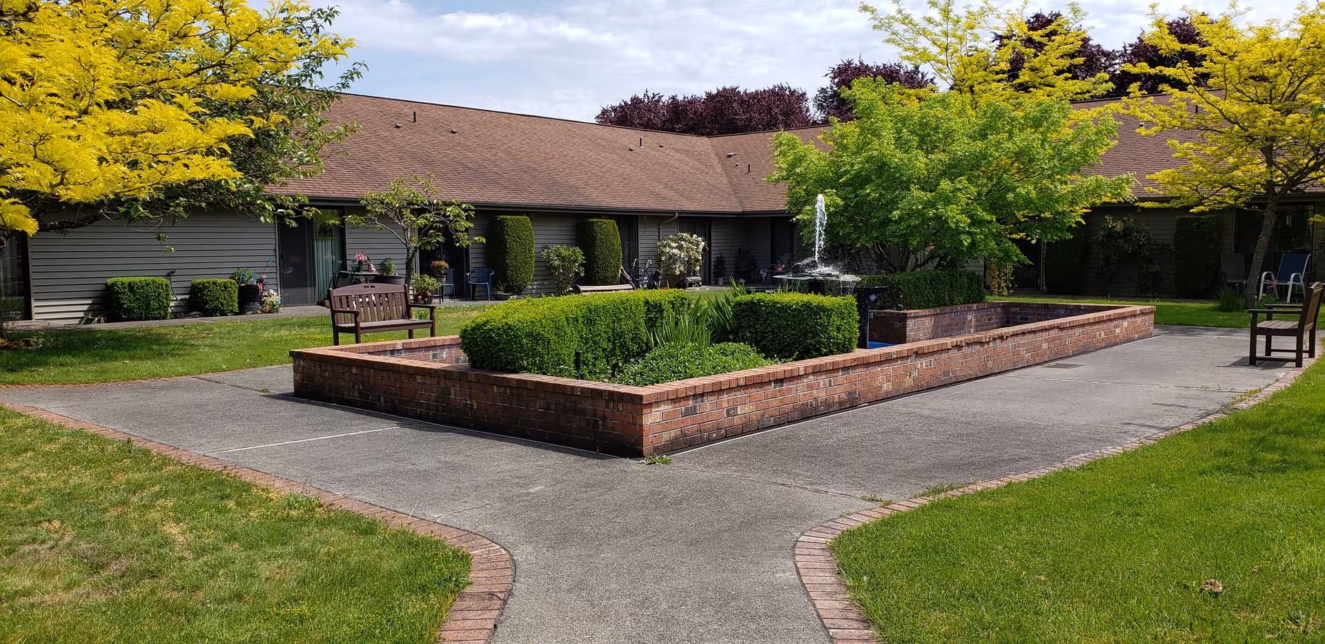 Sunlit courtyard with a central brick planter and fountain, benches, walkways, and a single-story residential building in the background.