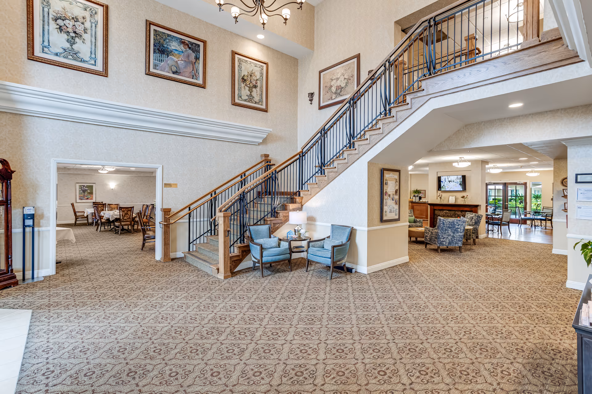 Interior view of a senior living facility featuring a carpeted lobby area with a staircase leading to an upper floor. There are two blue upholstered chairs with a small table and lamp between them at the base of the stairs. The walls are decorated with framed paintings. To the left, there is an entrance to a dining room with tables and chairs. To the right, there is a lounge area with armchairs and a fireplace, and beyond that, a sunlit seating area with large windows overlooking greenery outside.