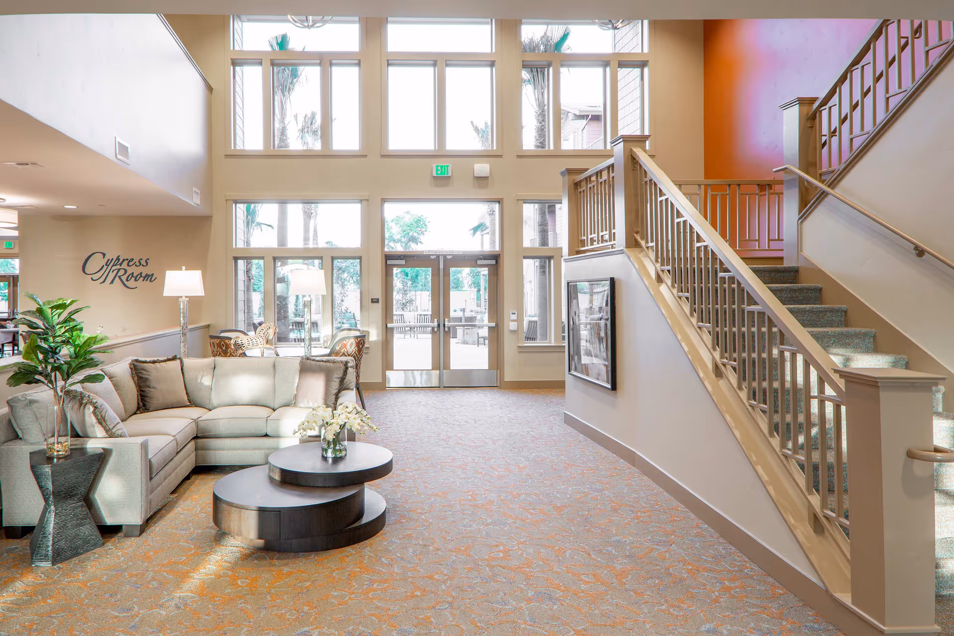 A bright and spacious interior common area with a large sectional sofa, two round coffee tables with a flower vase, a side table with a plant, and two floor lamps. There is a staircase with a wooden railing on the right side and large windows and glass doors at the back letting in natural light. The wall on the left has the words 'Cypress Room' written on it.