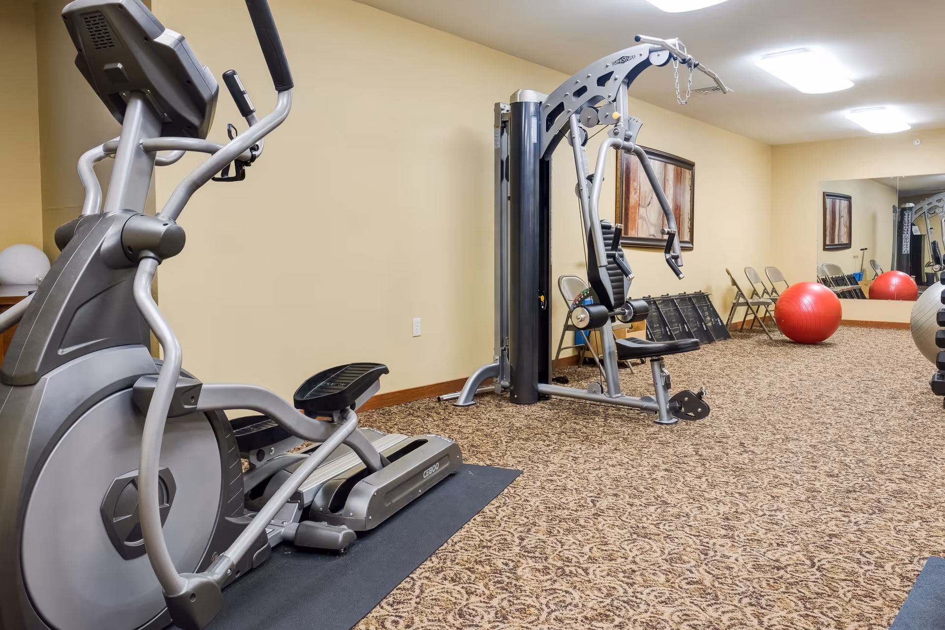 Interior view of a fitness room in a senior living facility with exercise equipment including an elliptical machine, a multi-function weight machine, and large red exercise balls. The room has patterned carpet, beige walls, a large mirror on one wall, and framed artwork.