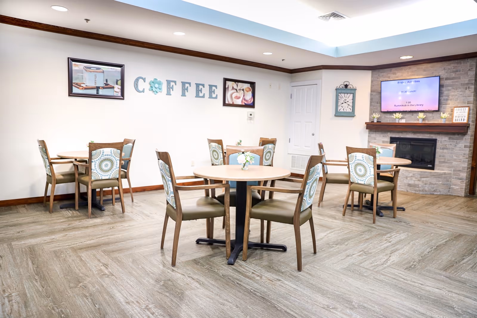 A bright and clean common area with round tables and chairs arranged for seating. The wall has decorative letters spelling 'COFFEE' with framed pictures above. There is a stone fireplace with a mounted TV displaying activities and a clock on the adjacent wall. The flooring is wood-patterned and the ceiling has recessed lighting.