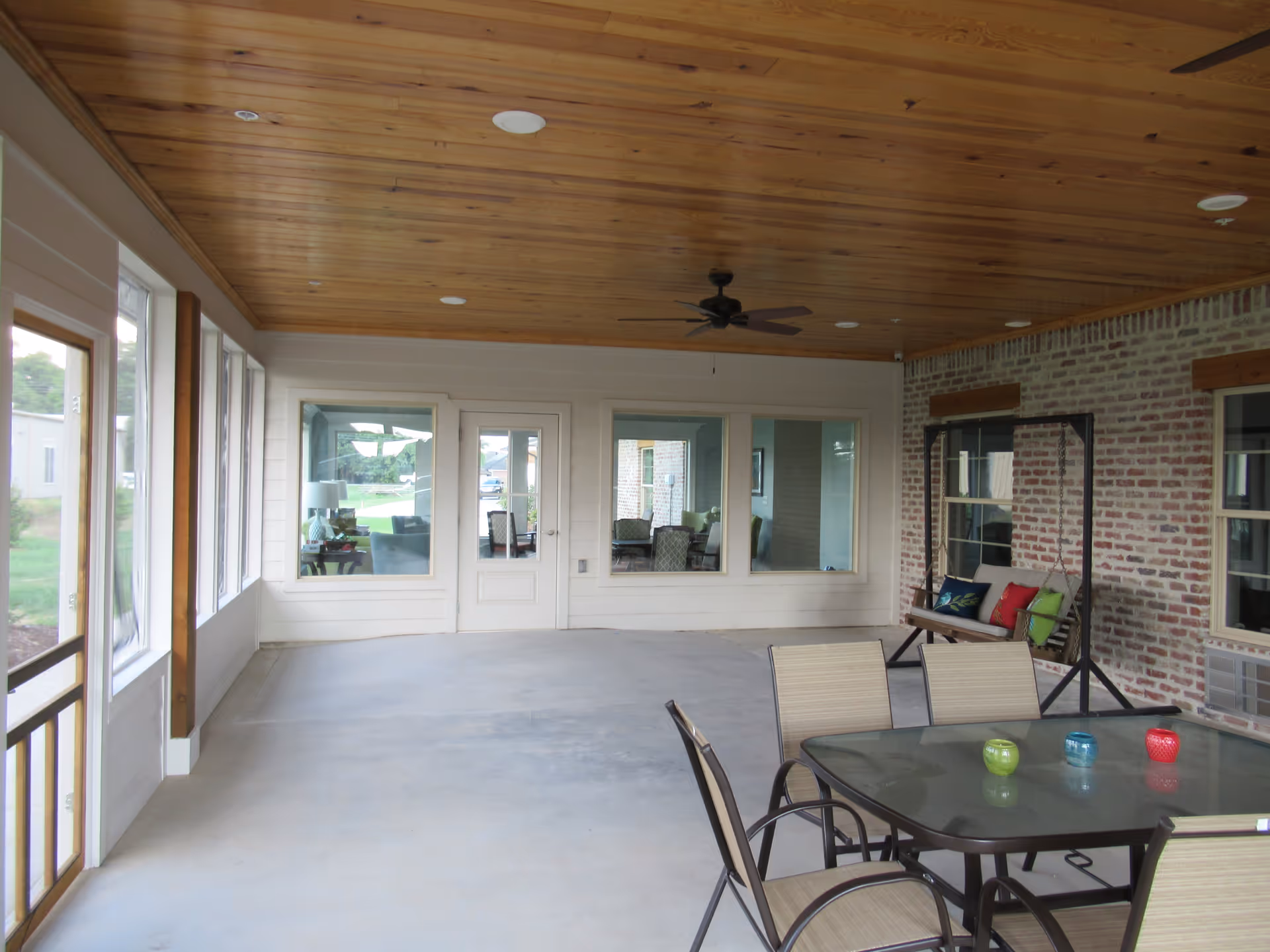 Covered outdoor patio area with a wooden ceiling, ceiling fan, brick wall, glass table with four chairs, and a wooden swing with colorful cushions. Large windows and a door look into an interior room with chairs and tables.