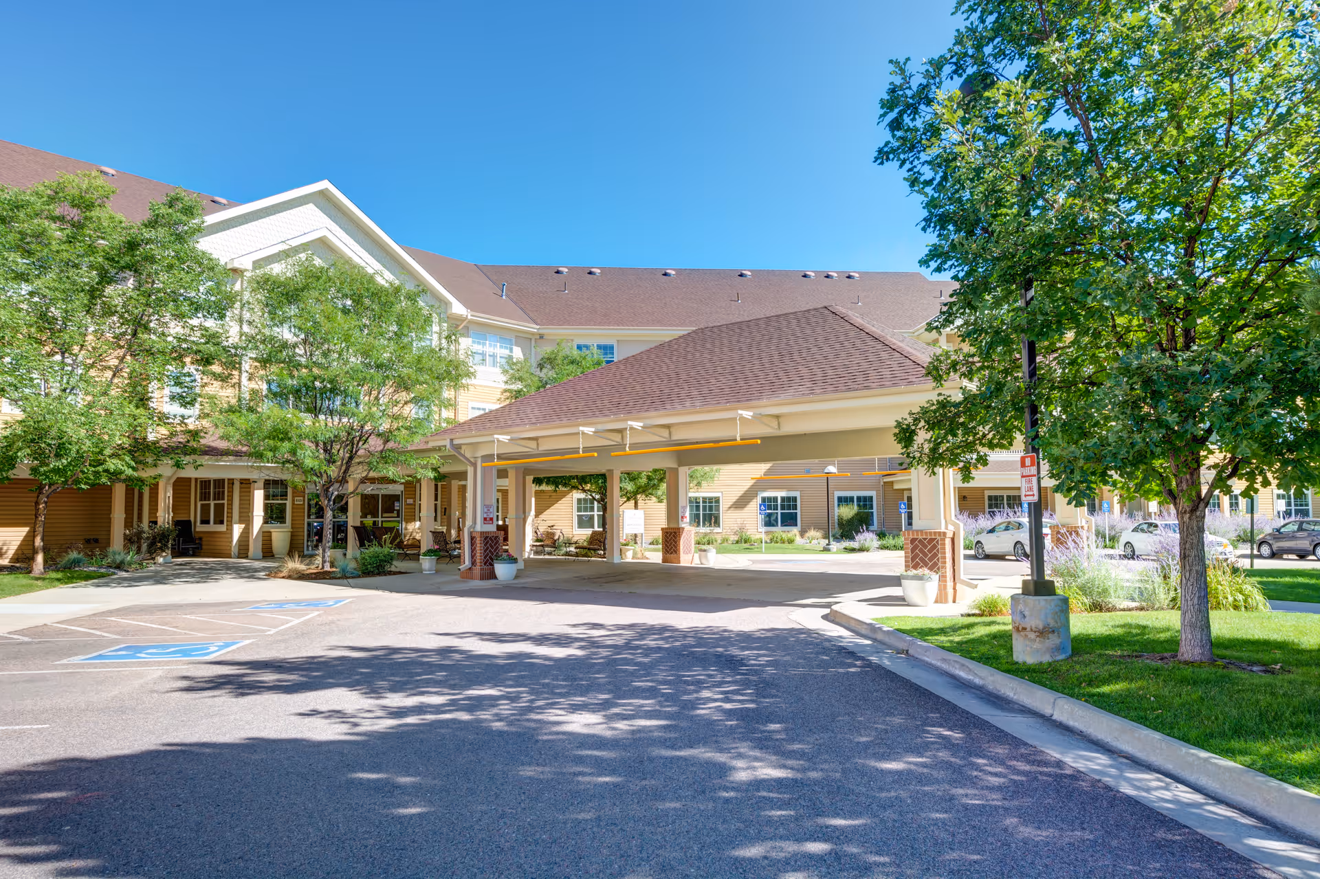 Front entrance and covered porte-cochere of a senior living building with trees, landscaping, and a parking area.