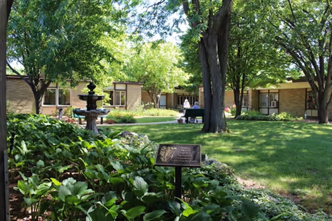 A peaceful outdoor garden area at a senior living facility with green grass, trees providing shade, a stone fountain, and a plaque on a stand surrounded by plants. In the background, there are single-story brick buildings with large windows and a person walking along a pathway.