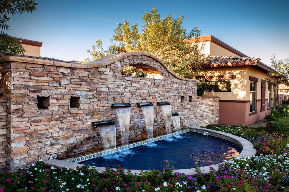 Outdoor stone water feature with four cascading waterfalls flowing into a small pool surrounded by colorful flowers and greenery, with a building in the background under a clear blue sky.