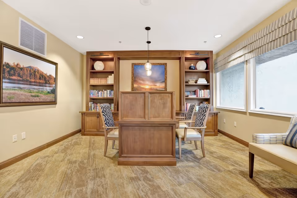 A cozy interior room with a wooden table and four upholstered chairs with patterned backs. Behind the table is a wooden built-in bookshelf filled with books and decorative items. A framed landscape painting hangs on the left wall, and two large windows with striped valances are on the right side, allowing natural light to enter. The room has beige walls and carpeted flooring.