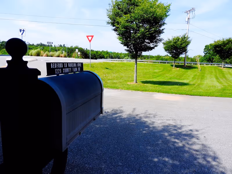 A black mailbox in the foreground with a sign that reads 'Bedford Co Nursing Home 1229 County Farm Rd' next to a paved road. There are green grassy areas with a few trees and road signs including a yield sign and a directional arrow sign in the background under a clear blue sky.