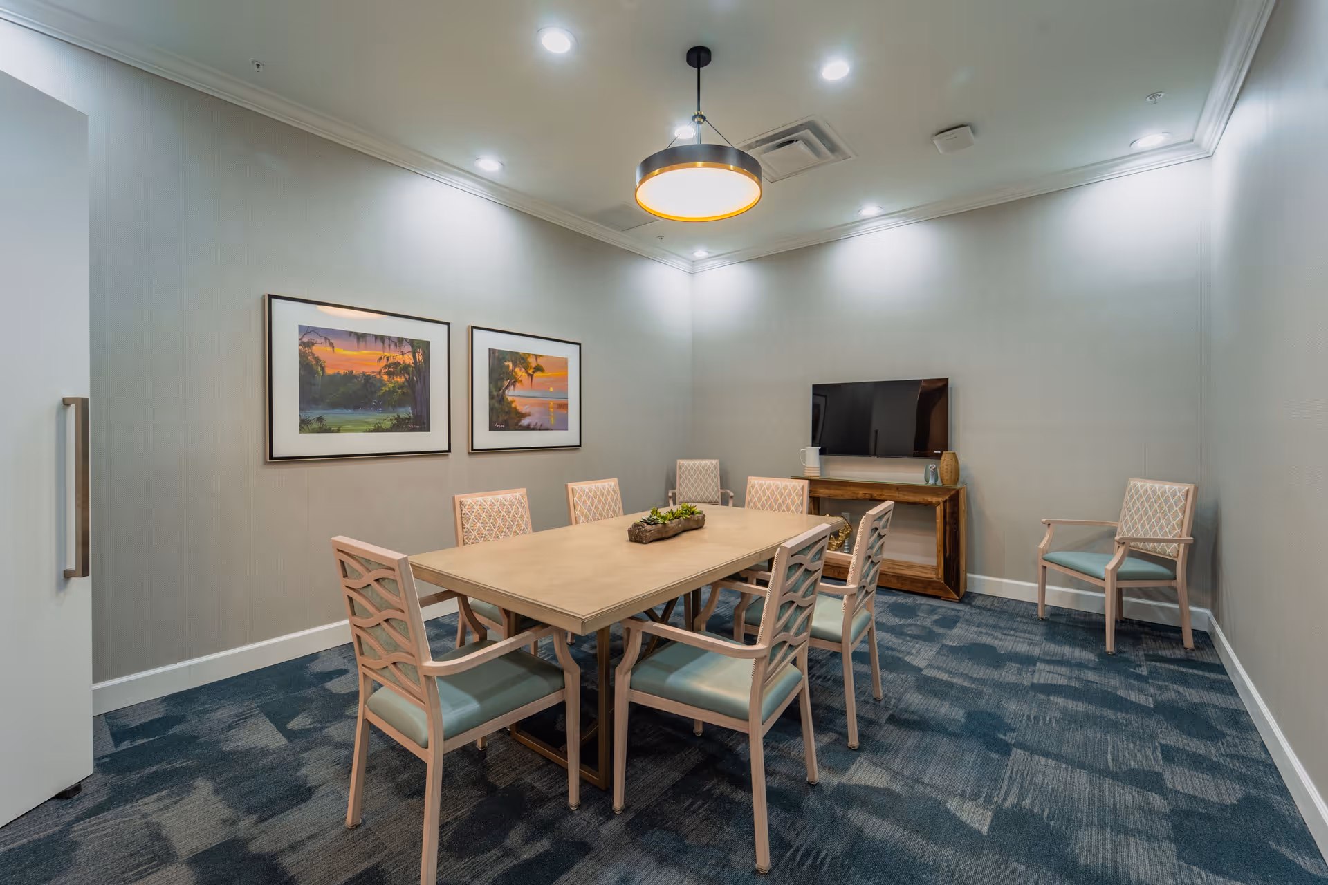 A small meeting or dining room with a rectangular wooden table surrounded by eight cushioned chairs. The room has light gray walls, two framed landscape paintings, a flat-screen TV on a wooden console, and a modern circular ceiling light fixture. The floor is carpeted in a blue patterned design.