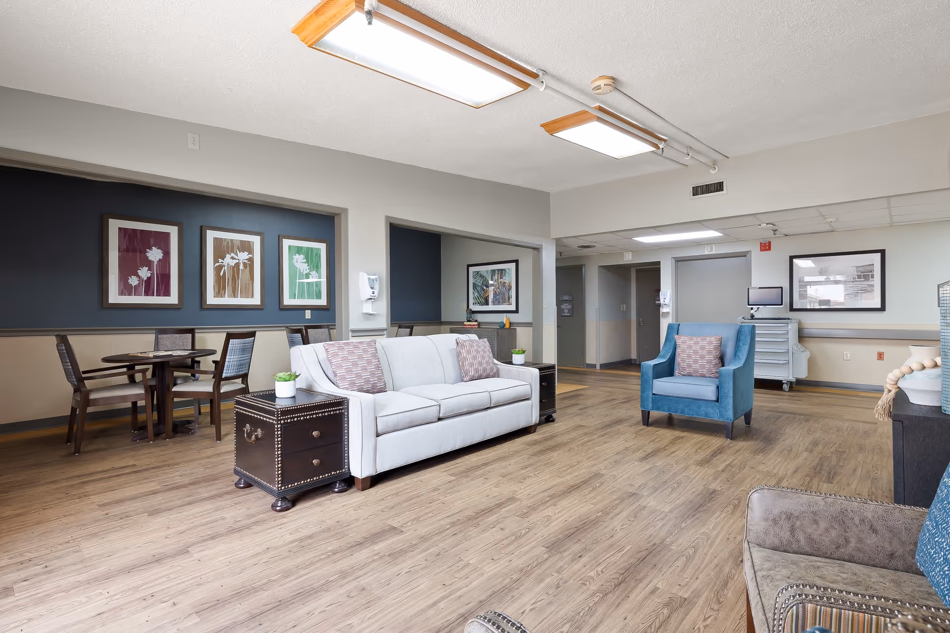 A spacious and well-lit common area in a senior living facility featuring a white sofa with patterned pillows, a blue armchair, a small round table with chairs, framed floral artwork on the walls, wood flooring, and overhead rectangular light fixtures.