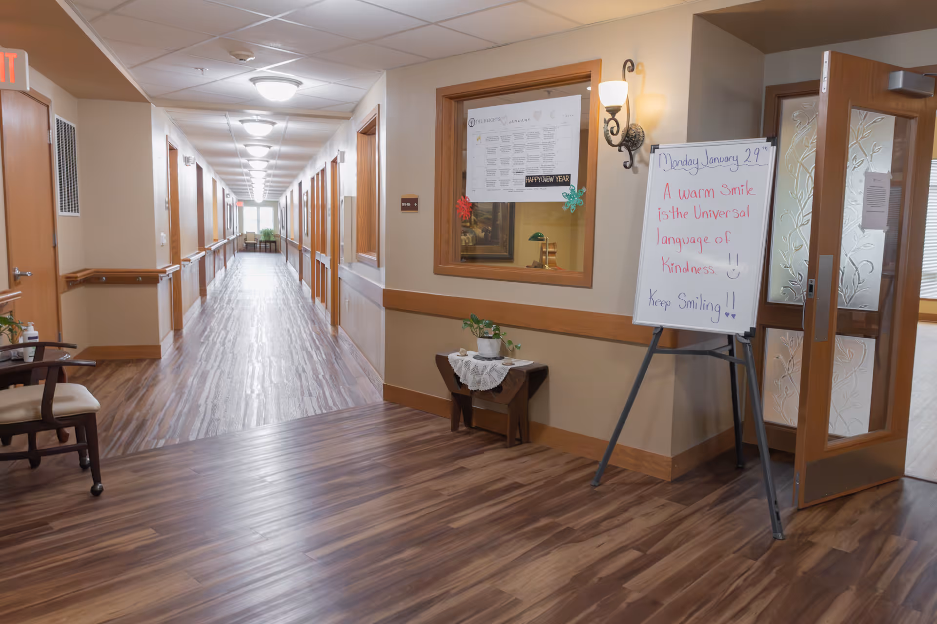 A clean, well-lit hallway in a senior living facility with wood flooring and beige walls. There are handrails along the walls and multiple closed doors on either side. At the end of the hallway, there is a window letting in natural light. To the right, there is a whiteboard on an easel with a motivational message, and a small table with a plant and a lace doily. A partially open door with frosted glass panels is also visible on the right.