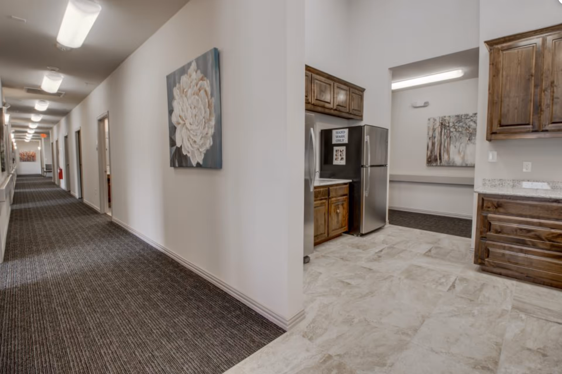 Interior corridor of an assisted living facility showing a long carpeted hallway with wall art and an adjacent kitchenette with wooden cabinets and a stainless refrigerator.