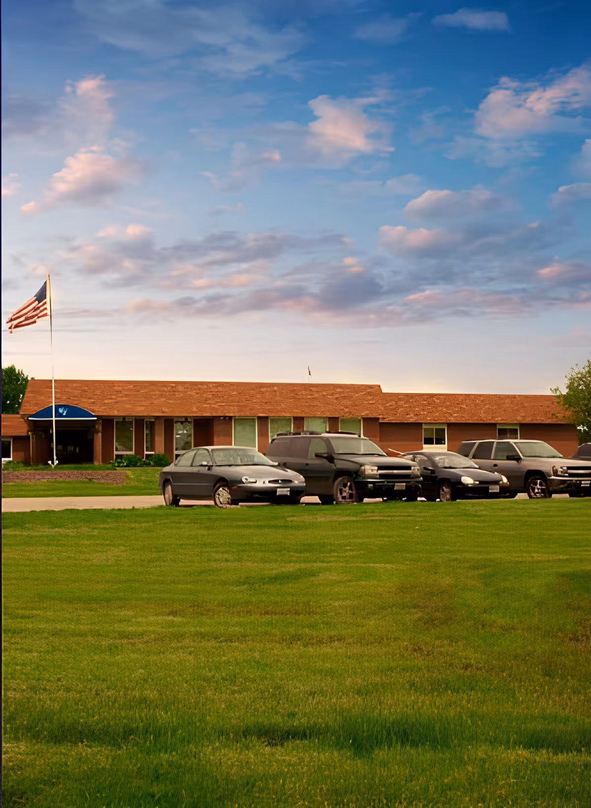 One-story senior living building with a row of parked cars, an American flag, and a large grassy lawn under a partly cloudy sky.