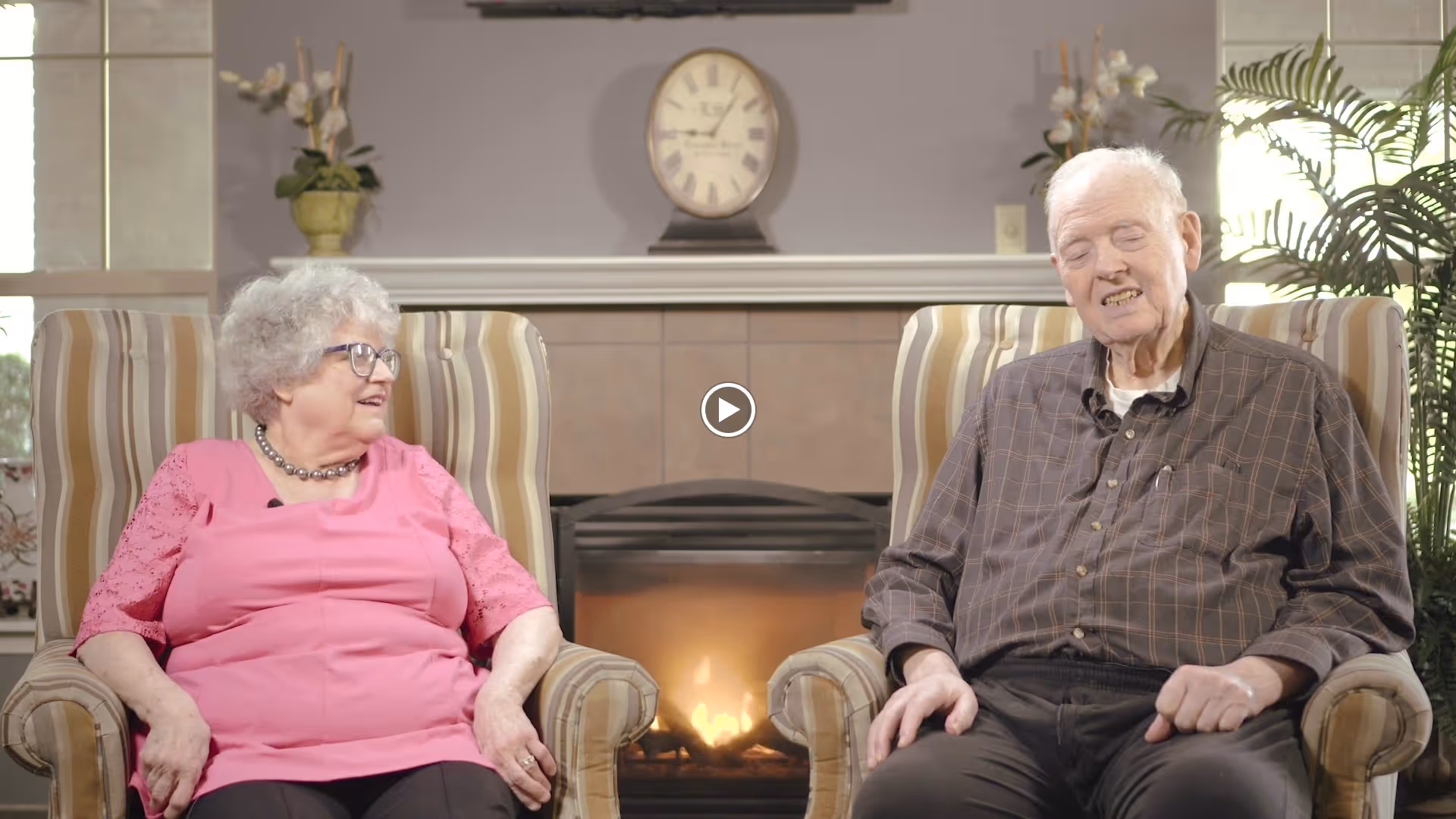 An elderly woman and an elderly man sitting in striped armchairs facing each other in a cozy living room with a lit fireplace and a clock on the mantel behind them. There are plants and decorative items on either side of the fireplace.