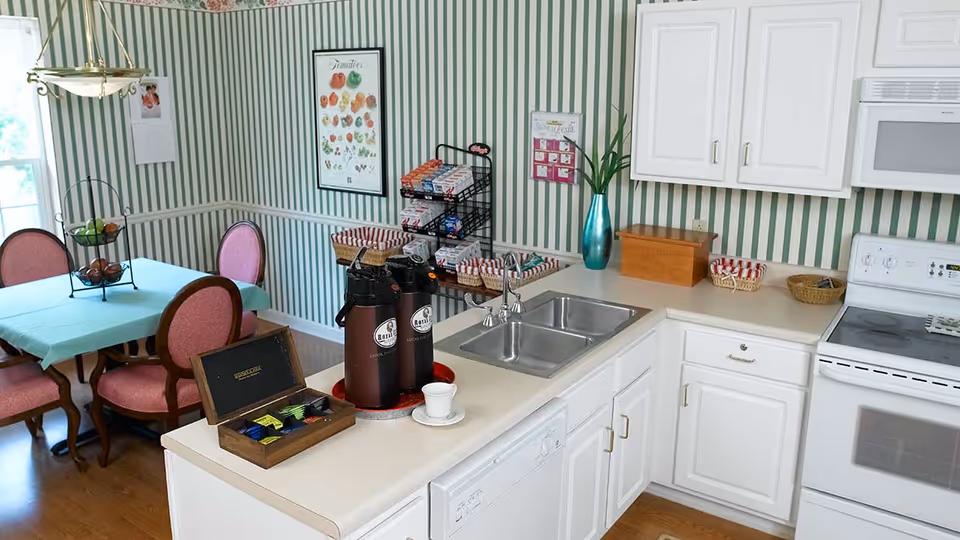 A kitchen area with white cabinets, a double sink, a white stove, and a microwave. On the countertop, there are two coffee dispensers, a wooden box with tea bags, and a white cup and saucer. In the background, there is a dining table with a turquoise tablecloth and four pink upholstered chairs. The walls have green and white vertical striped wallpaper, and there are framed pictures and a tall blue vase with a plant on the counter.