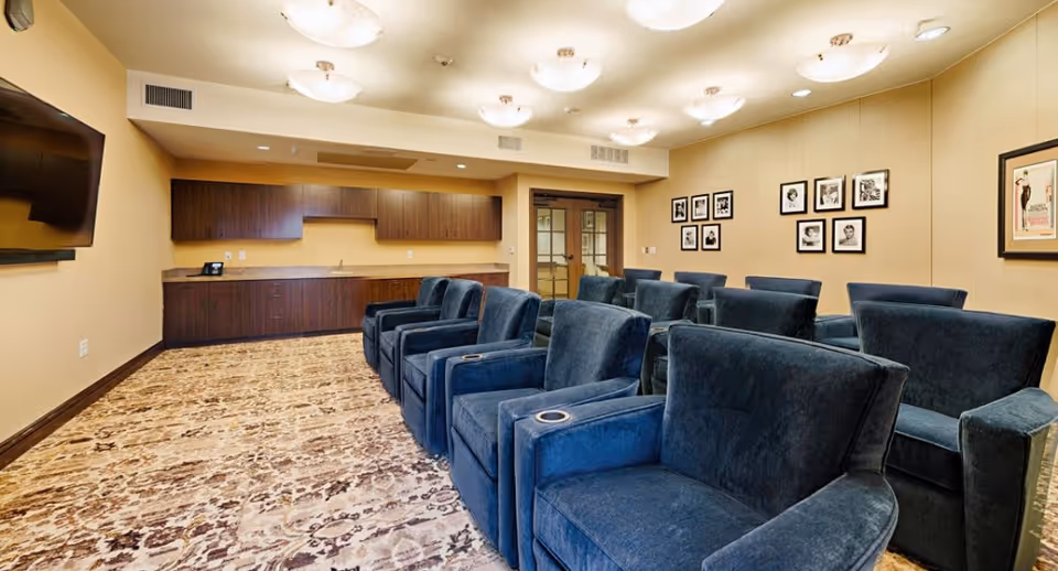 A cozy common room with multiple blue upholstered armchairs arranged in rows facing a wall-mounted TV. The room has beige walls adorned with framed black-and-white photographs and a framed poster. There is a long wooden cabinet with a countertop and a sink along one wall. The floor is covered with a patterned carpet, and the ceiling has several round light fixtures.