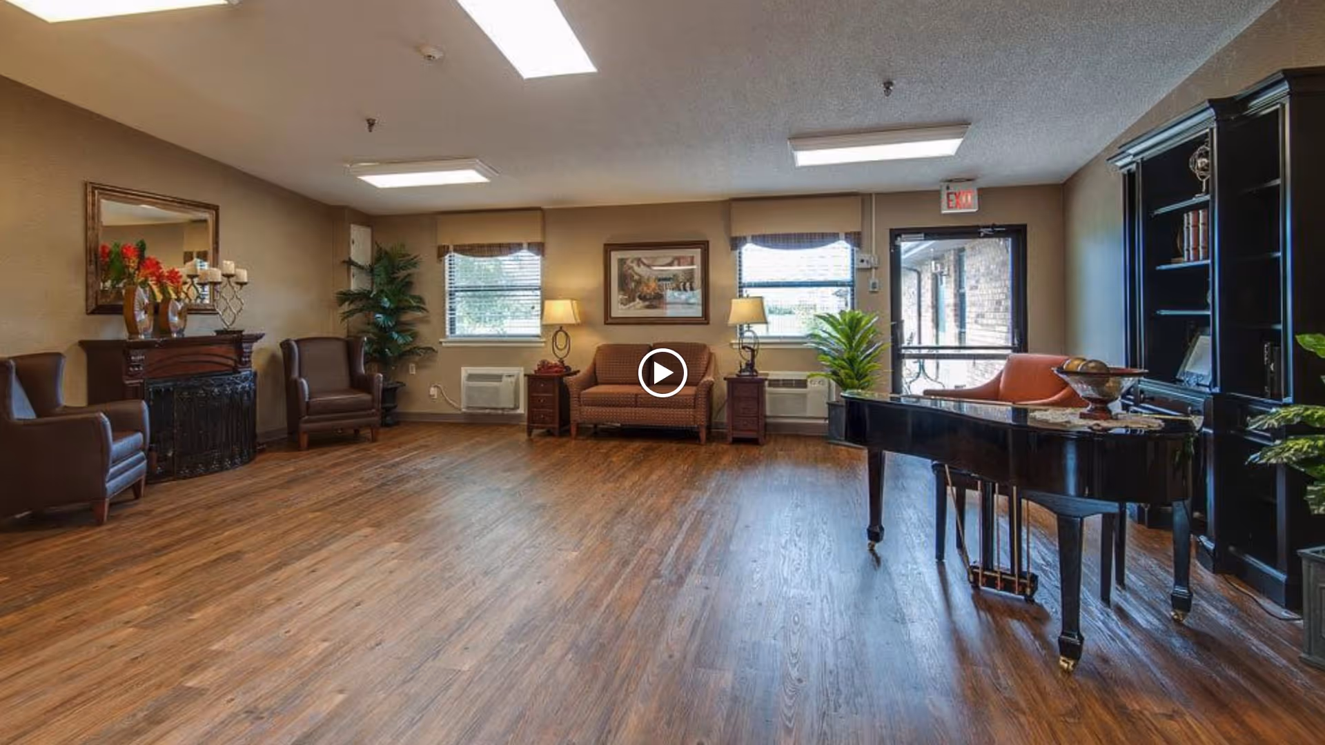 A spacious living room area with wooden flooring, featuring a black grand piano on the right, a black bookshelf, a brown sofa with two side tables and lamps, two brown armchairs near a decorative fireplace with a mirror above it, and several plants near the windows and door.
