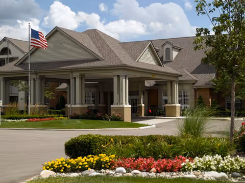 Exterior view of a senior living facility building with a covered entrance, an American flag on a flagpole, and well-maintained landscaping including colorful flowers and green shrubs under a partly cloudy sky.