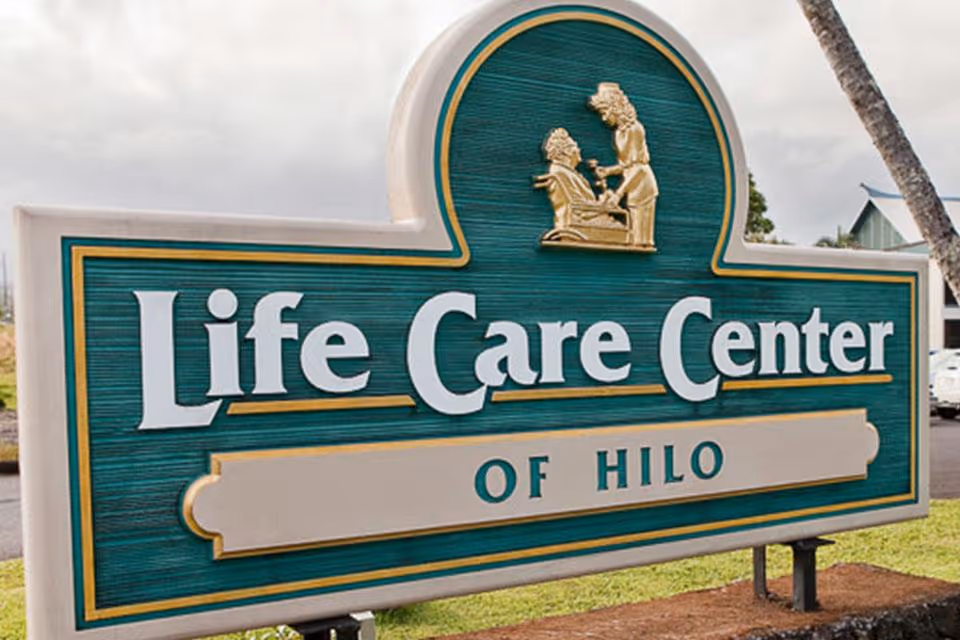 A green and gold sign for Life Care Center of Hilo with an emblem showing a caregiver assisting an elderly person.