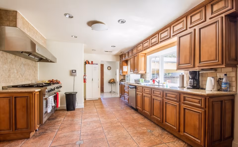 A spacious kitchen with wooden cabinets, a large window above the sink, a stainless steel stove with a range hood, a dishwasher, and various kitchen appliances on the countertops. The floor is tiled with brown tiles and the walls are light-colored.