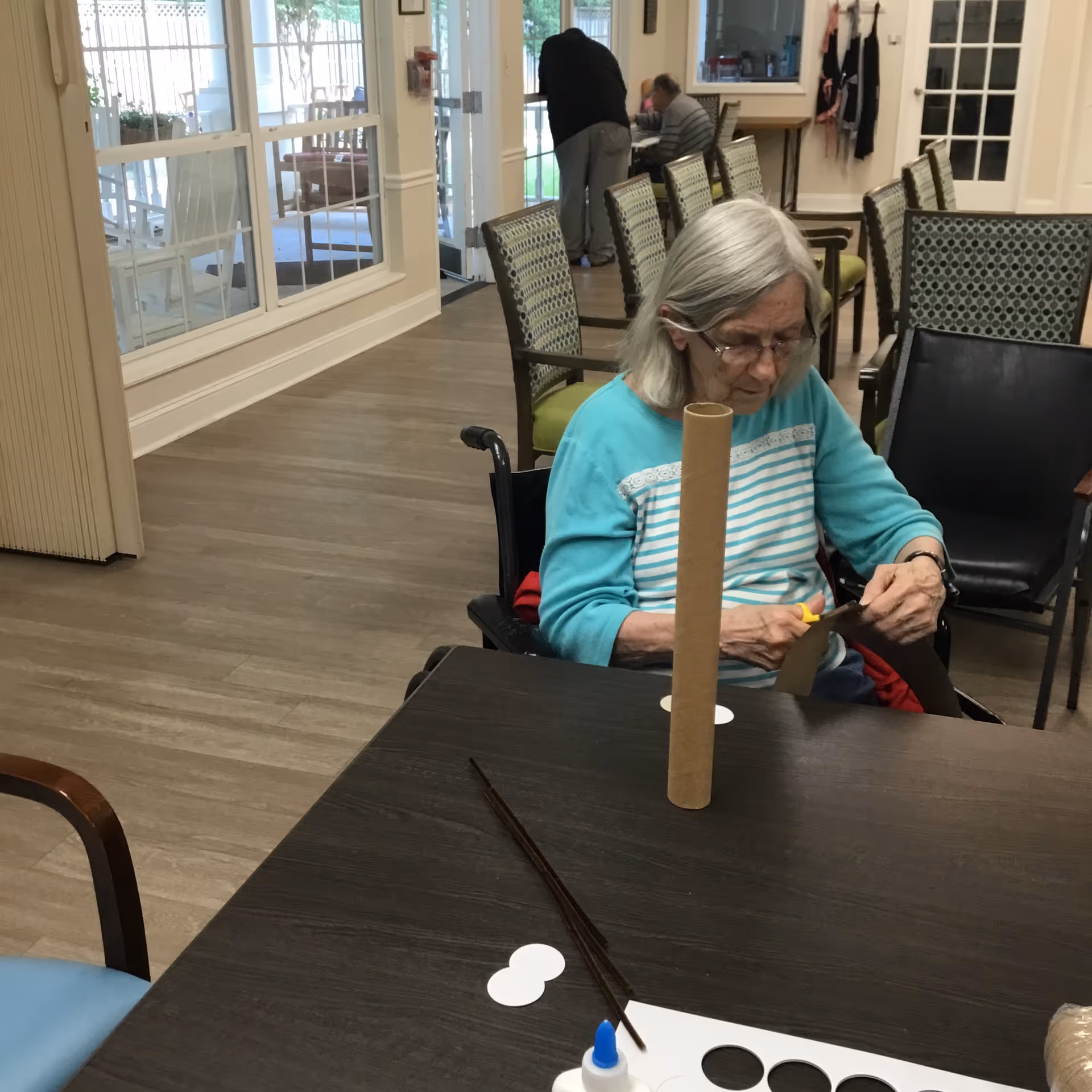 An elderly woman with gray hair and glasses sitting at a table in a common area, cutting a piece of brown paper with scissors. On the table are craft materials including a cardboard tube, glue, and paper cutouts. In the background, there are several chairs, a large window, and two other people engaged in activities.