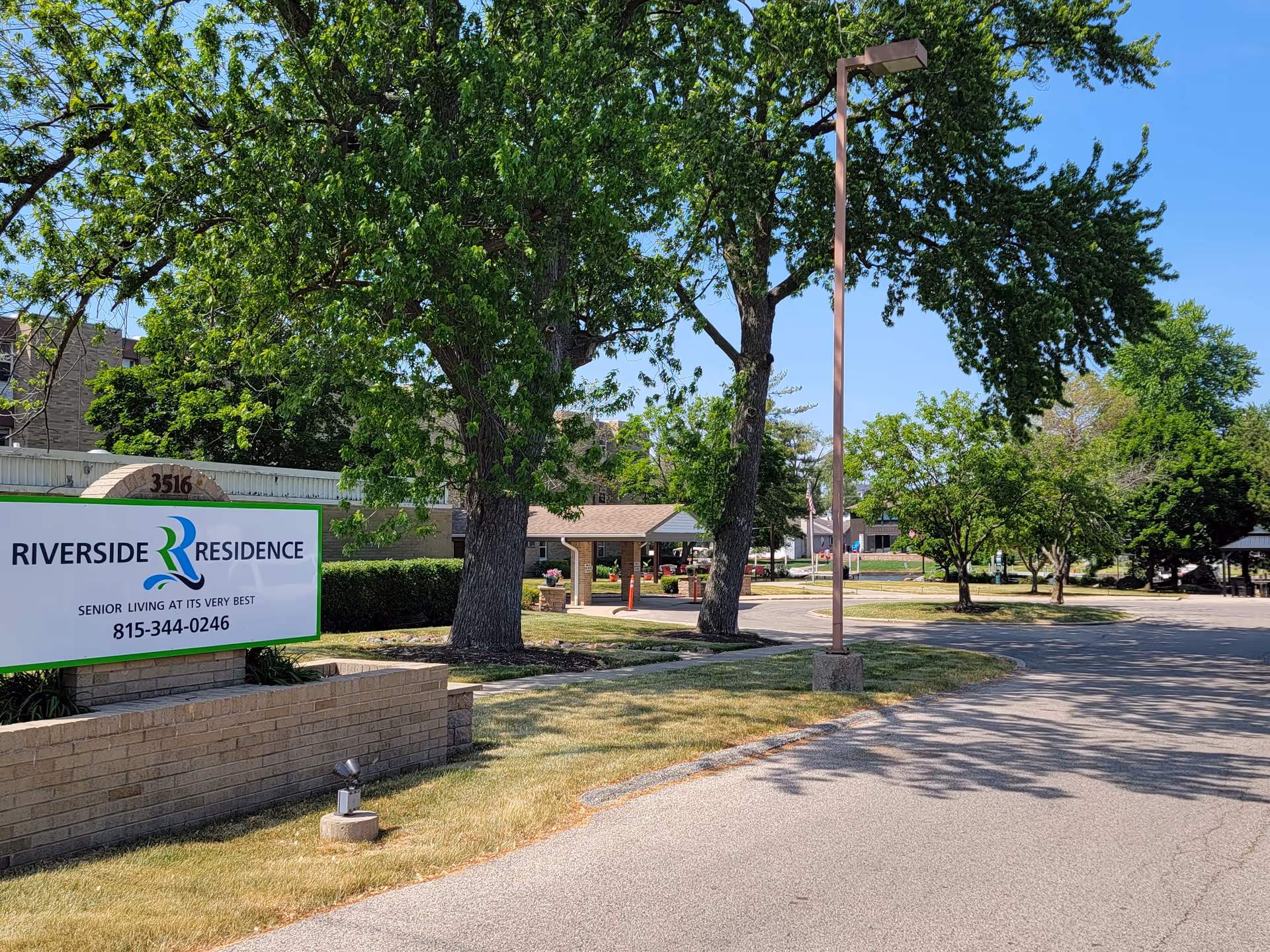 Outdoor view of Riverside Residence senior living facility with a large sign displaying the name and phone number. The scene includes a paved driveway, green trees, a street lamp, and a covered entrance area in the background under a clear blue sky.