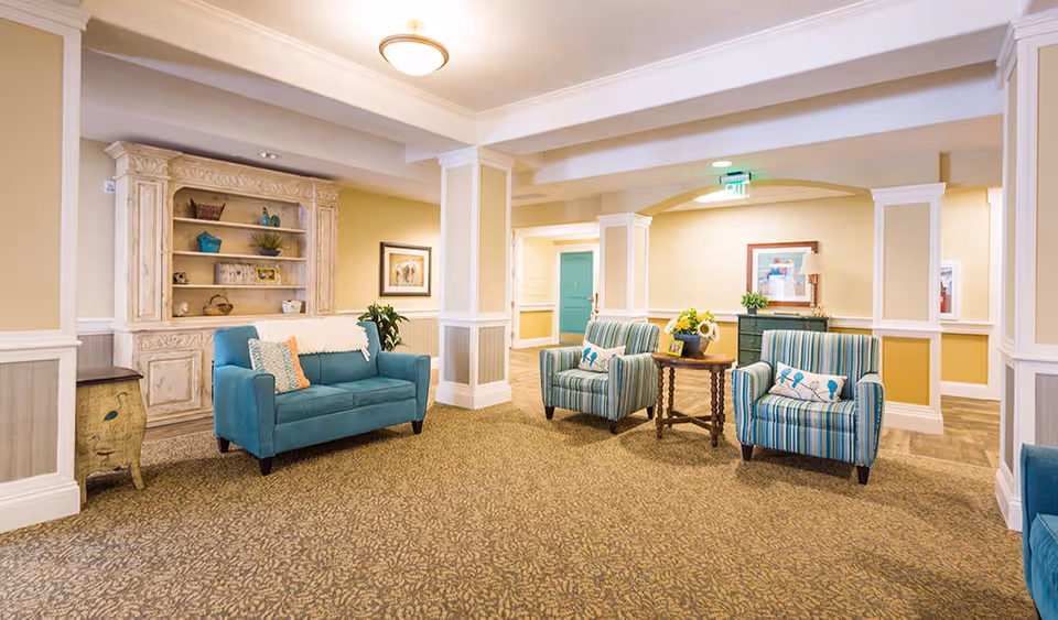 A bright and welcoming assisted living facility common area with beige walls and carpeted floor. The room features a teal loveseat with decorative pillows, two striped armchairs with a small wooden table between them holding a vase of sunflowers, and a vintage-style cream-colored cabinet with shelves displaying decorative items. The space is well-lit with ceiling lights and has white trim and columns.