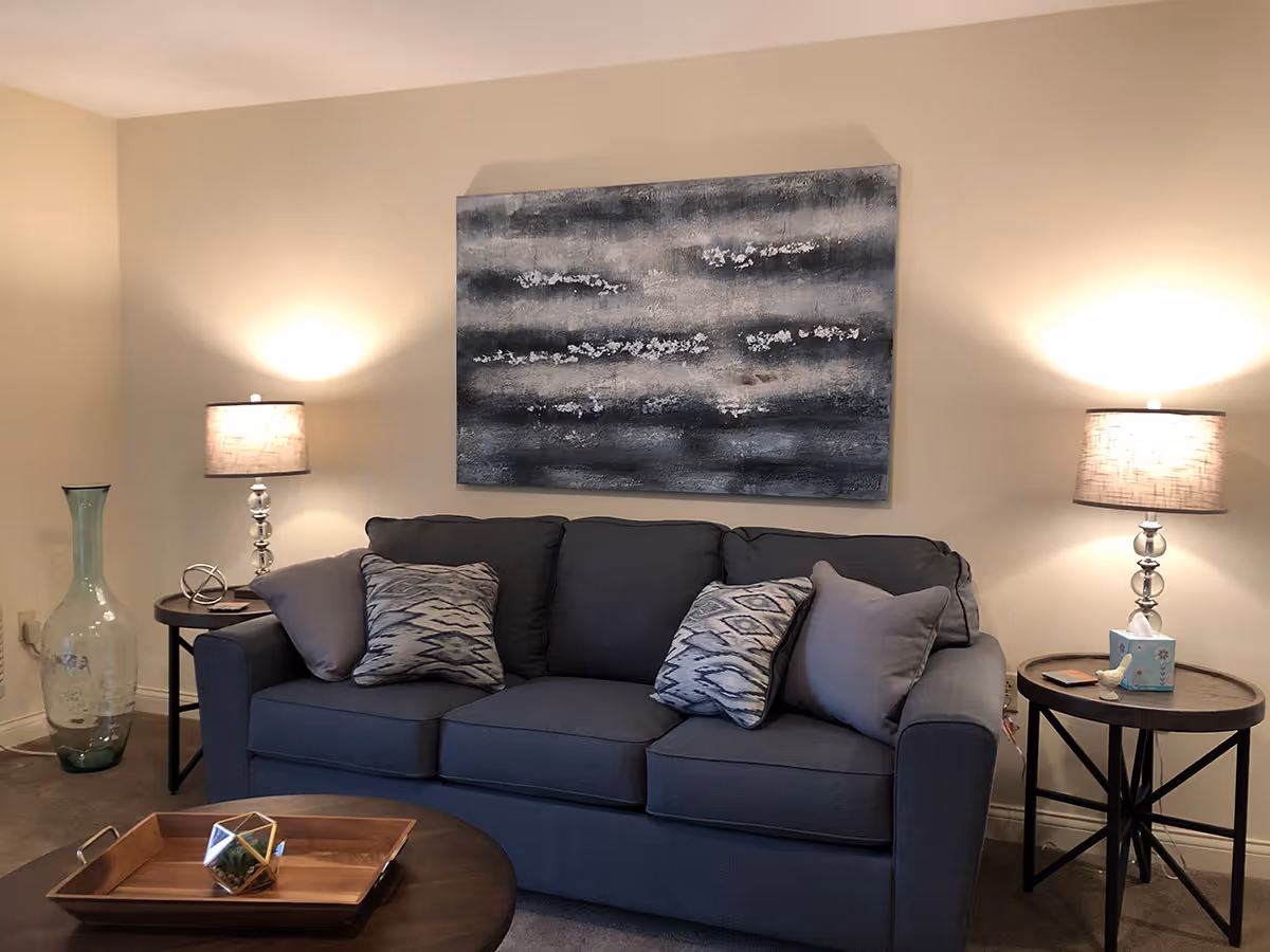 A cozy living room with a dark gray sofa adorned with four decorative pillows in shades of gray and patterned designs. On either side of the sofa are round wooden side tables, each with a lamp emitting warm light. Above the sofa hangs a large abstract painting in shades of gray, white, and black. A large glass vase is placed on the floor to the left of the sofa, and a wooden coffee table with a decorative tray and geometric terrarium is in the foreground.