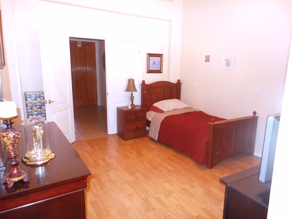 Simple bedroom with a wooden single bed, nightstand, dresser, lamp, and hardwood floor.