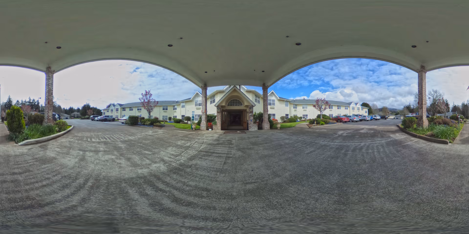 Covered driveway and main entrance of a two-story pale yellow senior living building with parked cars and landscaped grounds.