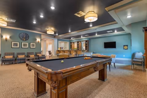 A spacious senior living common area featuring a pool table in the center, several chairs along the walls, a wall-mounted TV, and a dartboard. The room has a blue ceiling with recessed lighting and modern light fixtures, with a carpeted floor and light blue walls decorated with framed artwork and a large clock.