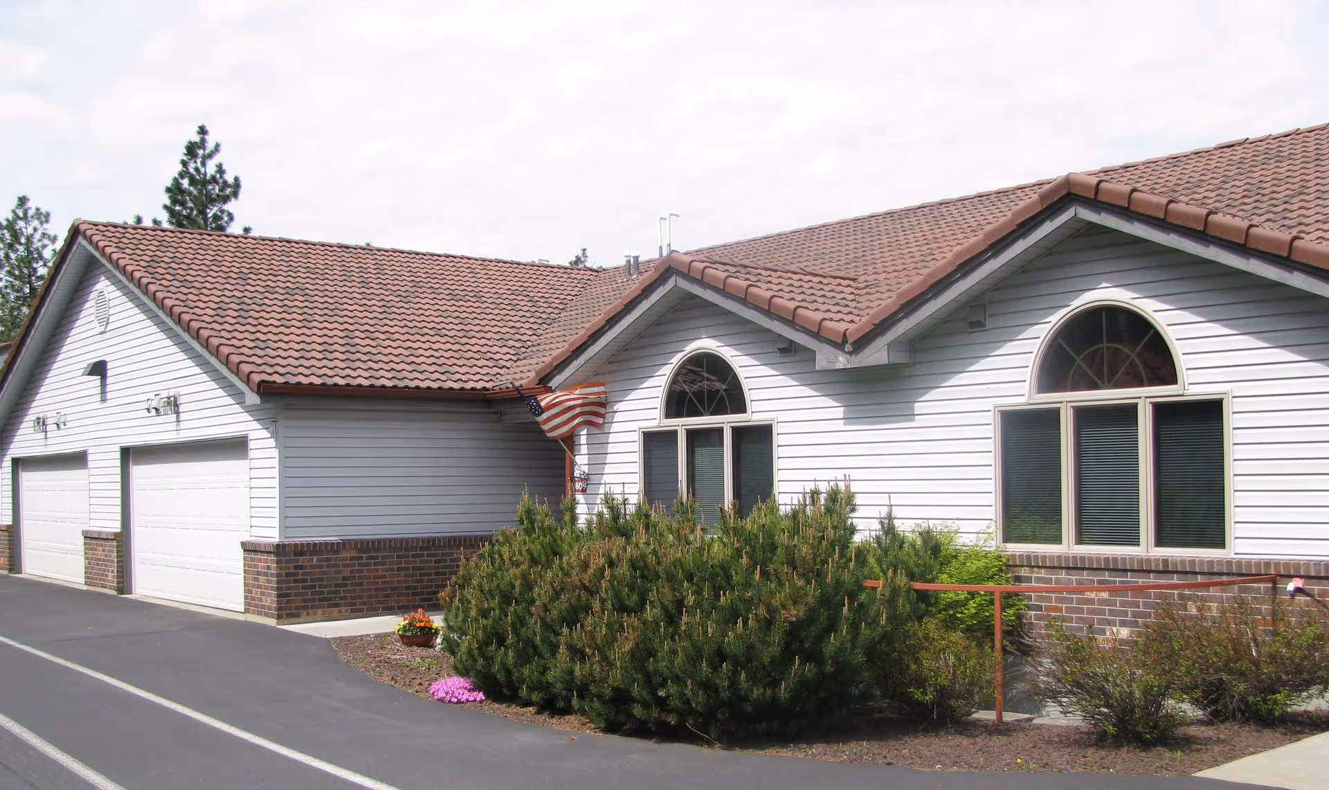 Front exterior of a single-story retirement building with white siding, arched windows, a red tile roof, garage doors, an American flag, and landscaping.