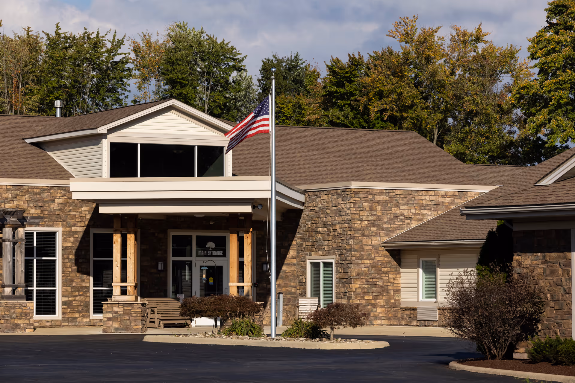 Front entrance of a stone-faced senior care building with an American flag on a pole and trees in the background.