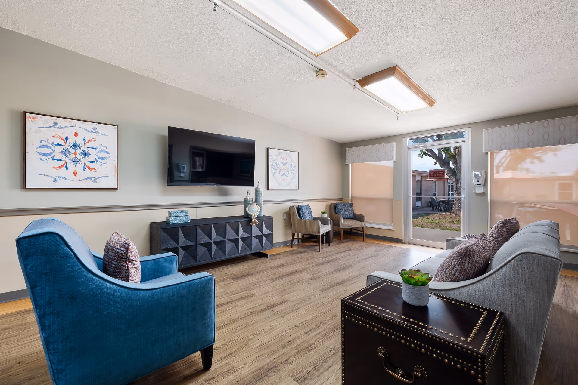 Sunlit common lounge with chairs, a sofa, wall-mounted TV, artwork, and wood-look flooring.