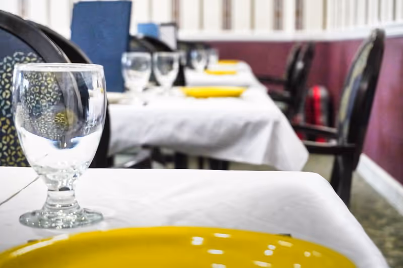 Close-up view of a dining table set with a clear water glass and a yellow plate, with additional tables and chairs visible in the background in a dining room setting.