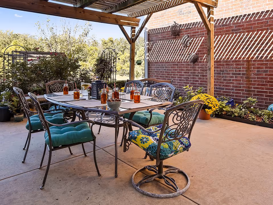 Outdoor patio area with a metal table and six chairs, some with green and patterned cushions. The table is set with several glasses of iced tea and a bowl of snacks. The patio is covered with a wooden pergola casting striped shadows on the brick wall and floor. There are plants and flowers around the patio, with greenery and trees visible in the background under a clear blue sky.