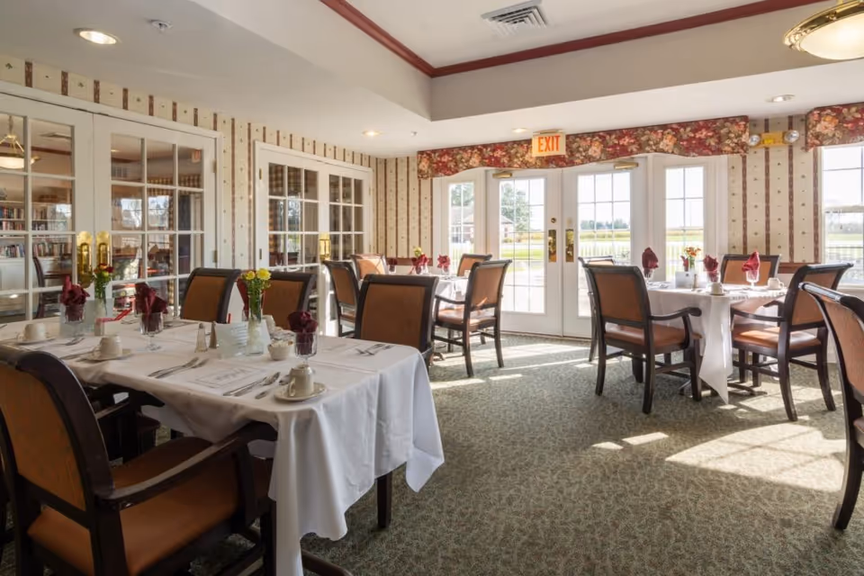 A bright dining room with several tables covered in white tablecloths, each set with cups, utensils, napkins, and small flower arrangements. The room has patterned wallpaper, carpeted floors, and large glass doors letting in natural light.