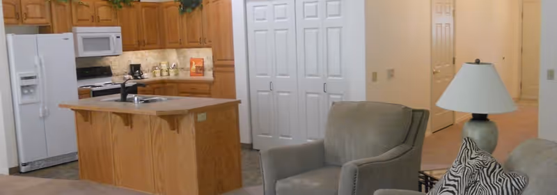 Interior view of a retirement community apartment showing a kitchen with wooden cabinets, a white refrigerator, a microwave, and a kitchen island with a sink. In the foreground, there is a living area with a gray armchair, a lamp on a side table, and a couch with patterned pillows. The background includes white double doors and a hallway.
