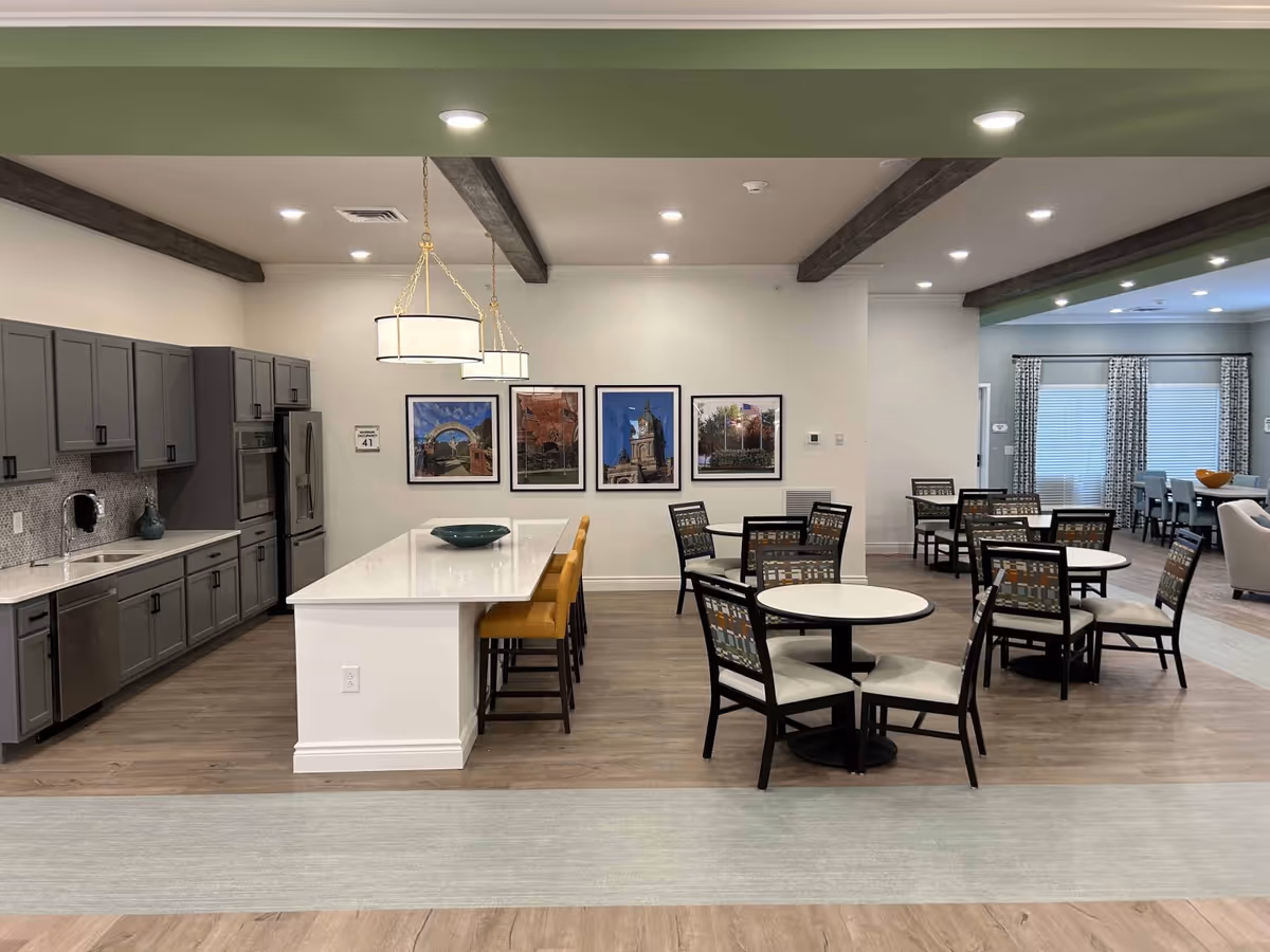 Open communal dining and kitchen area with a long island and bar stools, round dining tables and chairs, and framed artwork on the far wall.