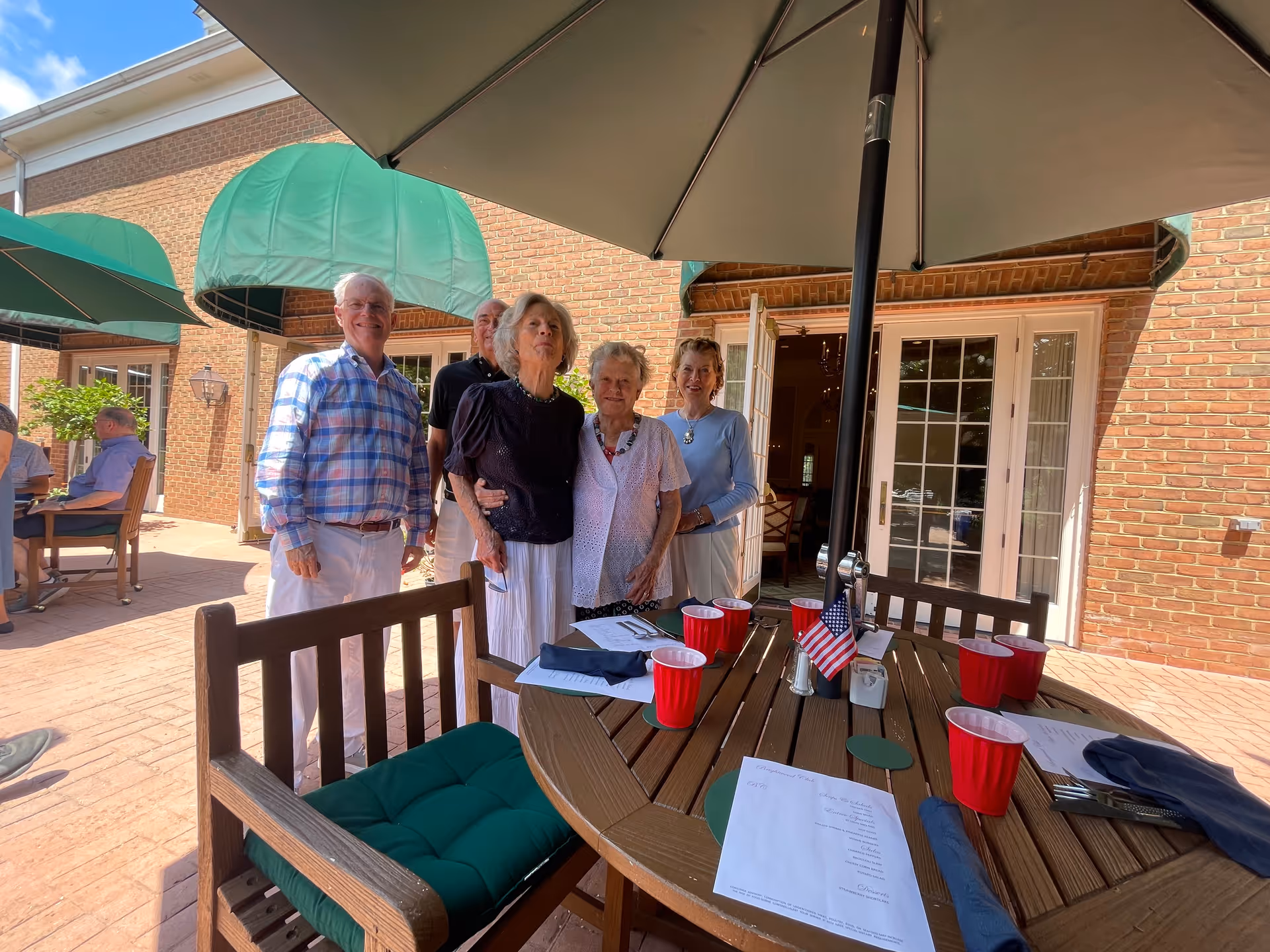 A group of five elderly people standing together and smiling on a patio outside a brick building with green awnings. In the foreground, there is a round wooden table with a large umbrella, red plastic cups, menus, and an American flag centerpiece. Other people are seated at tables in the background.