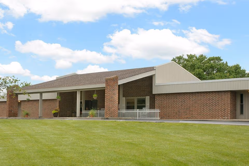 Exterior view of a single-story brick building with a sloped roof, a covered entrance supported by columns, hanging plants, and a small white picket fence in front. The building is surrounded by a well-maintained green lawn and trees under a partly cloudy blue sky.