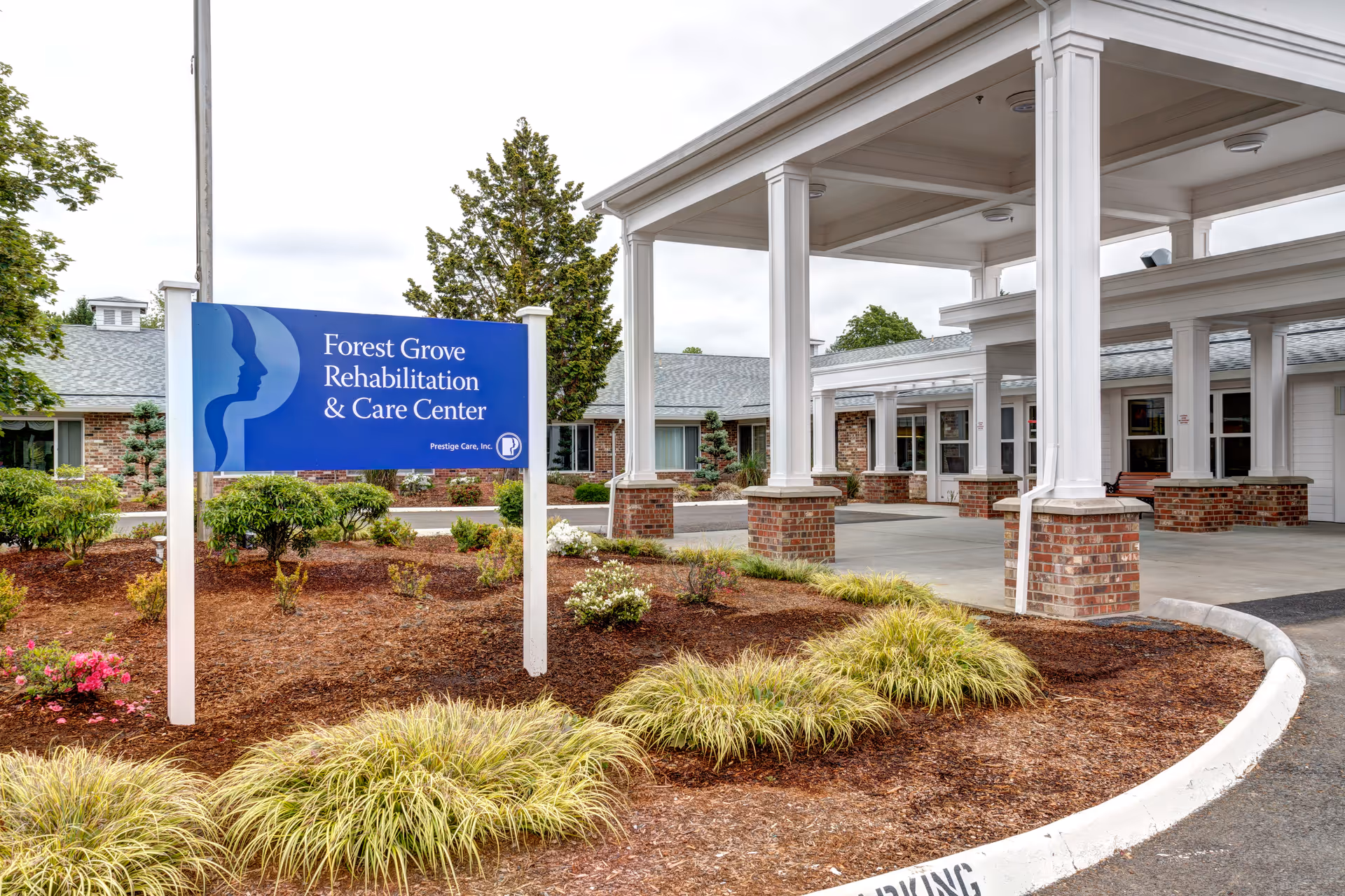Entrance area of Forest Grove Rehabilitation & Care Center showing a blue sign with the facility name, surrounded by landscaped bushes and plants, with a covered drop-off area supported by white columns and brick bases.