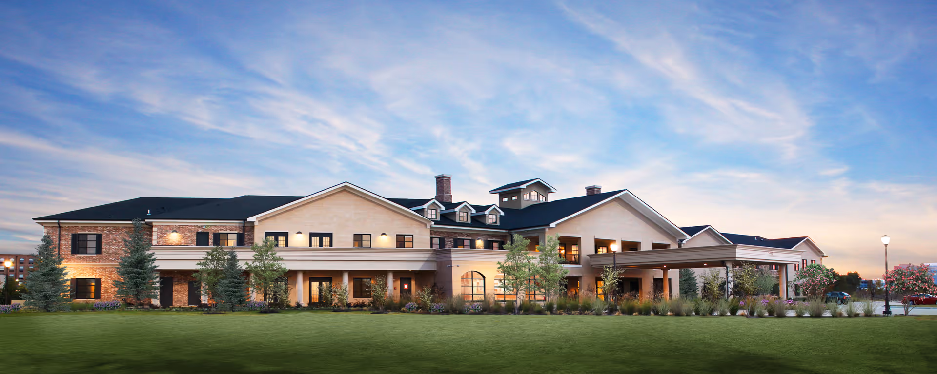 Exterior view of a large, two-story senior living facility building with a combination of brick and light-colored siding, surrounded by landscaped greenery and trees under a partly cloudy sky at dusk.