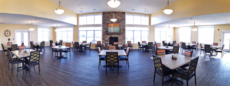 Spacious dining room with multiple square and round tables, each surrounded by chairs with patterned backs and striped cushions. The room features large windows allowing natural light to fill the space, a stone fireplace with colorful artwork above it, and light yellow walls with hanging ceiling lights. The floor is dark wood, and there is a clock on one wall near a door.