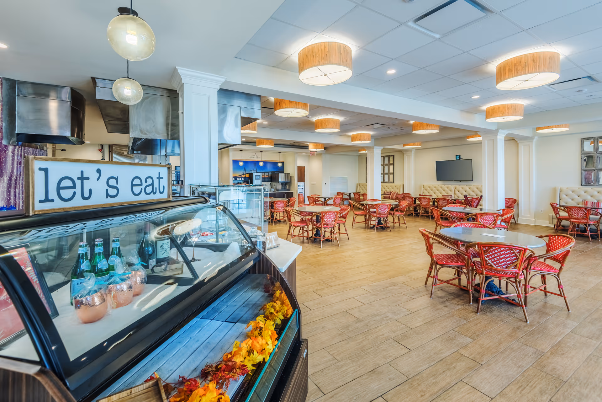 A spacious dining room with multiple round tables and red chairs arranged neatly. The room features warm lighting with circular ceiling fixtures and a large flat-screen TV mounted on the wall. In the foreground, there is a glass display case with beverages and a sign that reads 'let's eat'.