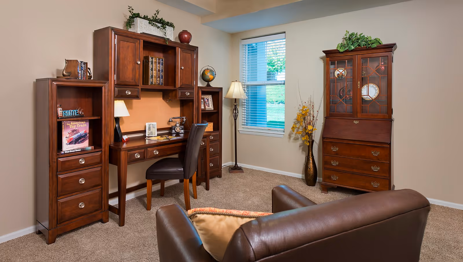 A cozy room with beige walls and carpet featuring a dark wooden desk with a hutch, a black leather chair, a tall wooden cabinet with glass doors displaying decorative plates, a floor lamp, a vase with yellow flowers, and a brown leather armchair with a beige pillow in the foreground.